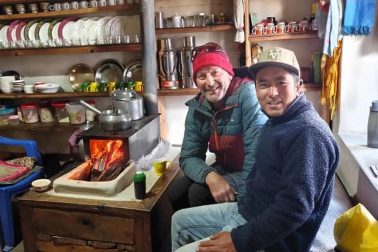 A trekker and local guide warming up by a traditional indoor stove in a mountain teahouse during Nepal trek tours.
