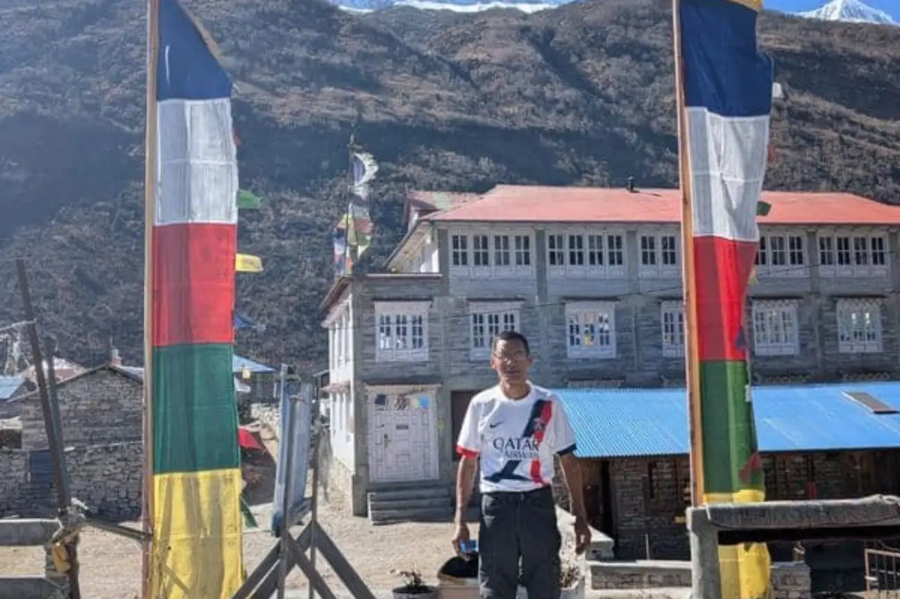 A person standing near tall prayer flags and mountain lodges during Nepal trek tours.